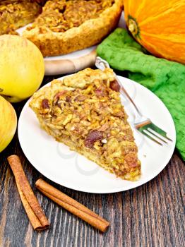 Piece of pie with apples, pumpkin, raisins and nuts in a white plate, cinnamon, napkin on a wooden board background