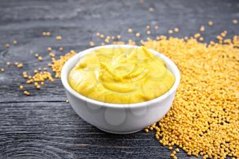 Mustard sauce in a white bowl and seeds on a black wooden board background