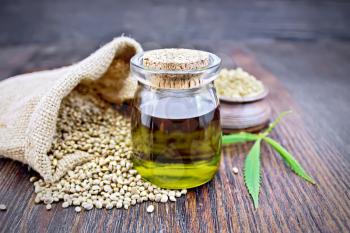 Hemp oil in a glass jar with flour in a clay bowl and grain in a bag, leaves and stalks of cannabis against a dark wooden board