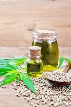 Hemp oil in two glass jars, a spoon with grains on a napkin of burlap, leaves and stalks of cannabis on a wooden board background