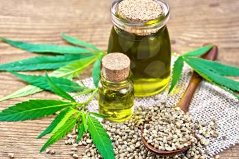 Hemp oil in two glass jars, a spoon with grains on sackcloth, leaves and stalks of cannabis against the background of an old wooden board