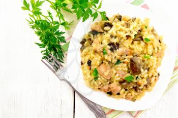 Rice risotto with mushrooms, chicken meat, cheese and garlic in a plate on a towel, fork and parsley on a wooden board background from above