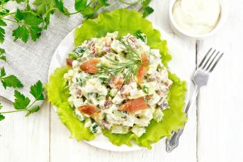 Salad from salmon, cucumber, eggs and avocado, dressed with mayonnaise on lettuce leaves in a plate, towel, dill, parsley and fork on a light wooden background from above