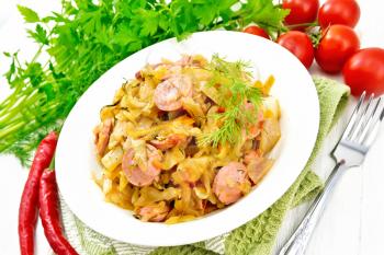 Cabbage stew with sausages in a white plate on a kitchen towel, tomatoes, parsley and fork on a wooden board background