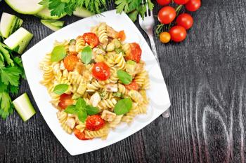 Fusilli with chicken, zucchini and tomatoes in a plate, fork, basil and parsley on wooden board background from above