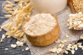 Oatmeal flour in bowl, milk in jug, oatmeal in spoon on napkin of burlap, grain in bag, oaten stalks against wooden board
