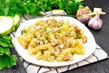 Fusilli pasta with chicken breast, zucchini, cream and pine nuts in a plate on napkin, garlic, fork and parsley on a dark wooden board background