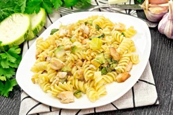 Fusilli pasta with chicken breast, zucchini, cream and pine nuts in a white plate on towel, garlic, fork and parsley on wooden board background