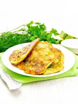 Zucchini fritters, dill and parsley in a plate on a napkin, sour cream in platter on white wooden board background
