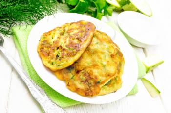 Zucchini fritters, dill and parsley in a plate on a kitchen towel, sour cream in saucer on wooden board background
