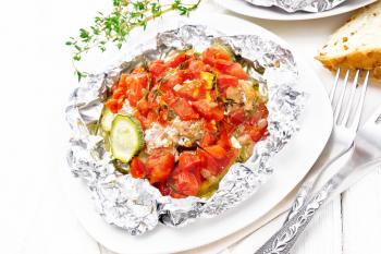 Pink salmon with zucchini, tomatoes, onions, garlic and thyme, baked in foil on a plate, towel, fork and bread on white wooden board background