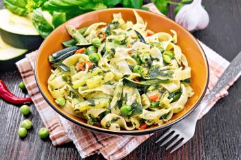 Tagliatelle pasta with zucchini, green peas, asparagus beans, hot peppers and spinach in kitchen towel, garlic, fork and basil on wooden board background