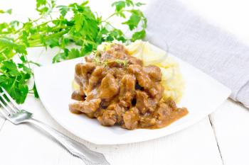 Beef goulash in tomato sauce with mashed potatoes in a plate, towel, parsley and fork on background of light wooden board