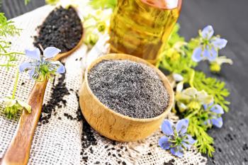 Flour of black caraway in a bowl, seeds in a spoon burlap, oil in bottle and twigs Nigella sativa with blue flowers and green leaves on dark wooden board background