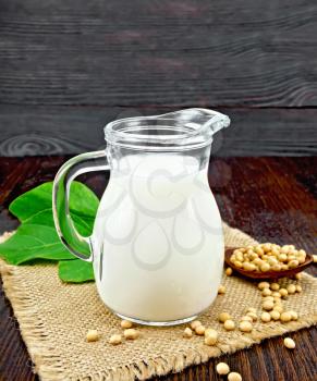 Soy milk in a jug, green leaf, soybeans in a spoon and burlap on the background of wooden boards