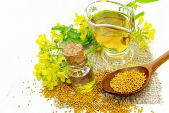 Mustard oil in a glass bottle and gravy boat, grains in a spoon and burlap, yellow mustard flowers on light wooden board background