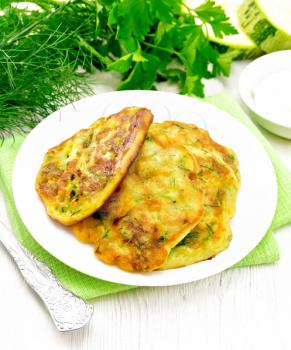 Fritters of zucchini, dill and parsley in a plate on a towel, sour cream in saucer on the background of light wooden board