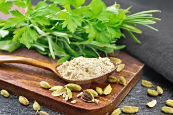 Ground cardamom in a spoon, seasoning capsules, napkin, fresh parsley and rosemary on black wooden board background
