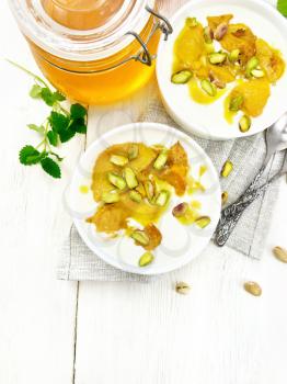 Dessert of yogurt, persimmon and honey with vanilla, cardamom and pistachios in two bowls on a napkin, mint and spoon on wooden board background from above
