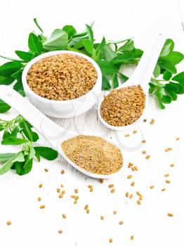 Ground fenugreek in a spoon, spice seeds in a bowl and spoon with green leaves on a background of light wooden board