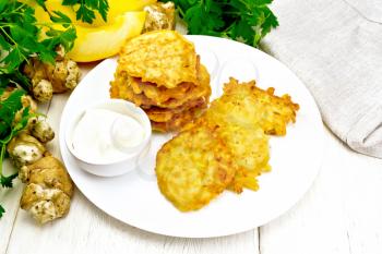Pumpkin and Jerusalem artichoke fritters with sour cream in a plate, napkin, parsley on light wooden board background