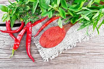 Ground hot red pepper in a spoon on burlap, chili pods and spicy greens on wooden board background from above