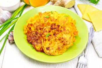 Fritters of pumpkin and cheese with ginger in a green plate, sour cream in a bowl, napkin, parsley and fork on background of light wooden board
