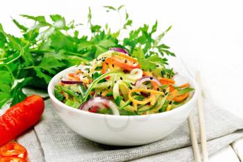 Spicy salad of cucumbers, carrots, chili peppers, purple onions, cilantro and black sesame seeds, seasoned with vinegar and lemon juice in a bowl on a towel on wooden board background