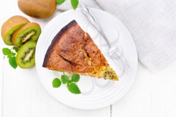 Piece of sweet cake with kiwi and honey, mint and a fork in a plate, a napkin on a wooden board background from above