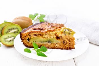 A piece of sweet cake with kiwi and honey, mint and fork in a plate, towel on a wooden board background
