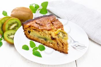 Piece of sweet cake with kiwi and honey, mint and fork in a plate, napkin on a wooden board background