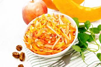 Pumpkin, carrot and apple salad with pecans seasoned with vegetable oil in a bowl on a napkin, mint on wooden board background