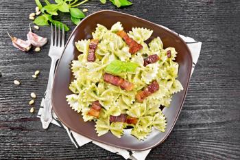 Farfalle pasta with pesto sauce, fried bacon and basil in a plate on a towel against dark wooden board on top