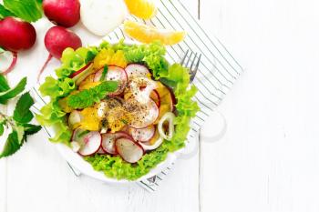 Radish, onion and orange salad with mint, vegetable oil and spices on lettuce in a plate on a napkin on wooden board background from above