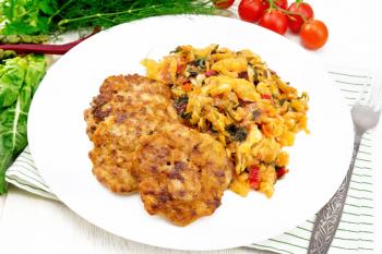 Fritters of minced meat with stewed cabbage in a plate, fork on towel, tomatoes, parsley and chard on light wooden board background
