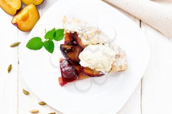 Piece of sweet pie with plum, sugar, cardamom and ice cream in a plate, sprigs of green mint, a napkin on wooden board background from above