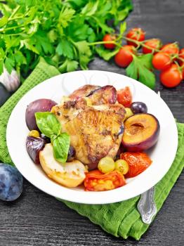 Baked chicken with tomatoes, apples, plums and grapes in a plate on a napkin, garlic, parsley and basil on dark wooden board background