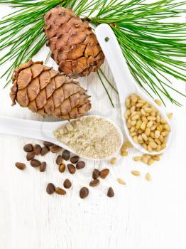 Cedar flour and peeled nuts in two spoons, two cones, a green cedar branch on wooden board background from above