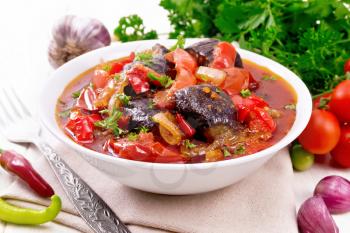 Vegetable ragout with eggplant, tomatoes, bell peppers, onions and spices in a plate on towel, garlic, parsley, hot peppers and a fork on wooden board background