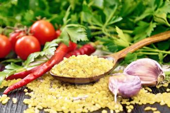 Bulgur groats - steamed wheat grains - in a spoon on sacking, tomatoes, hot peppers, garlic and parsley on the background of a dark wooden board