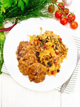 Fritters of minced meat with stewed cabbage in a plate, a fork on towel, tomatoes, parsley and chard on background of light wooden board from above