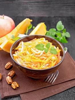 Pumpkin, carrot and apple salad with pecans seasoned with vegetable oil in a bowl on a napkin, mint on black wooden board background