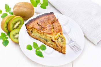 Piece of sweet cake with kiwi and honey, mint and a fork in a plate, a napkin on a white wooden board background