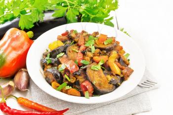 Vegetable ragout with eggplant, tomatoes, sweet and hot peppers, onions, carrots, fried with herbs and spices in plate on a towel, garlic, parsley on the background of light wooden board