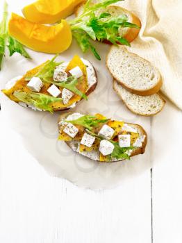 Bruschetta with baked pumpkin, salted feta cheese, ricotta, arugula and spices on parchment, napkin and vegetable slices on wooden board background from above