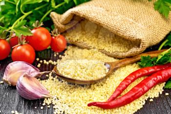Raw couscous in a spoon and a sack of burlap, tomatoes, hot peppers, herbs and garlic on wooden board background
