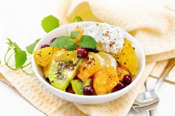 Fruit salad with banana, orange, kiwi, cranberries and baked pumpkin, whipped cream, sprinkled with chocolate and coconut with mint in a bowl on towel on a wooden board background