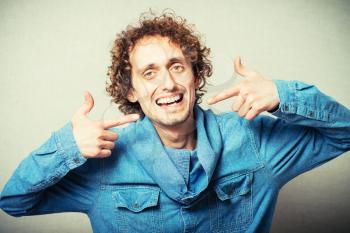 Curly young man laughs showing forefinger on the teeth after the dentist. On a gray background
