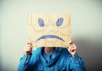 Curly man with a kraft cardboard instead of a head, a sad smiley. On a gray background.