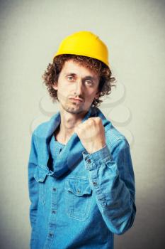 Curly man in a yellow helmet shows the evil revenge, blow fist. On a gray background.